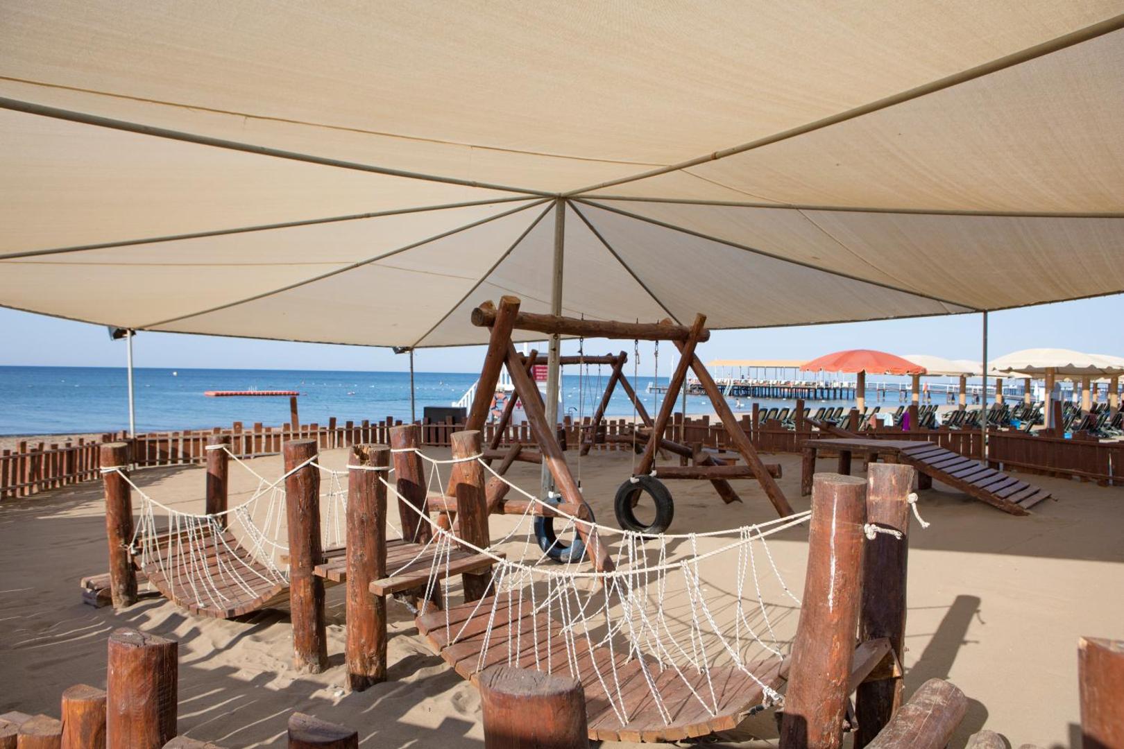 Pine Beach Belek bērnu rotaļu laukums pludmales smiltīs zem nojumes ar koka konstrukcijām un šūpolēm pie jūras