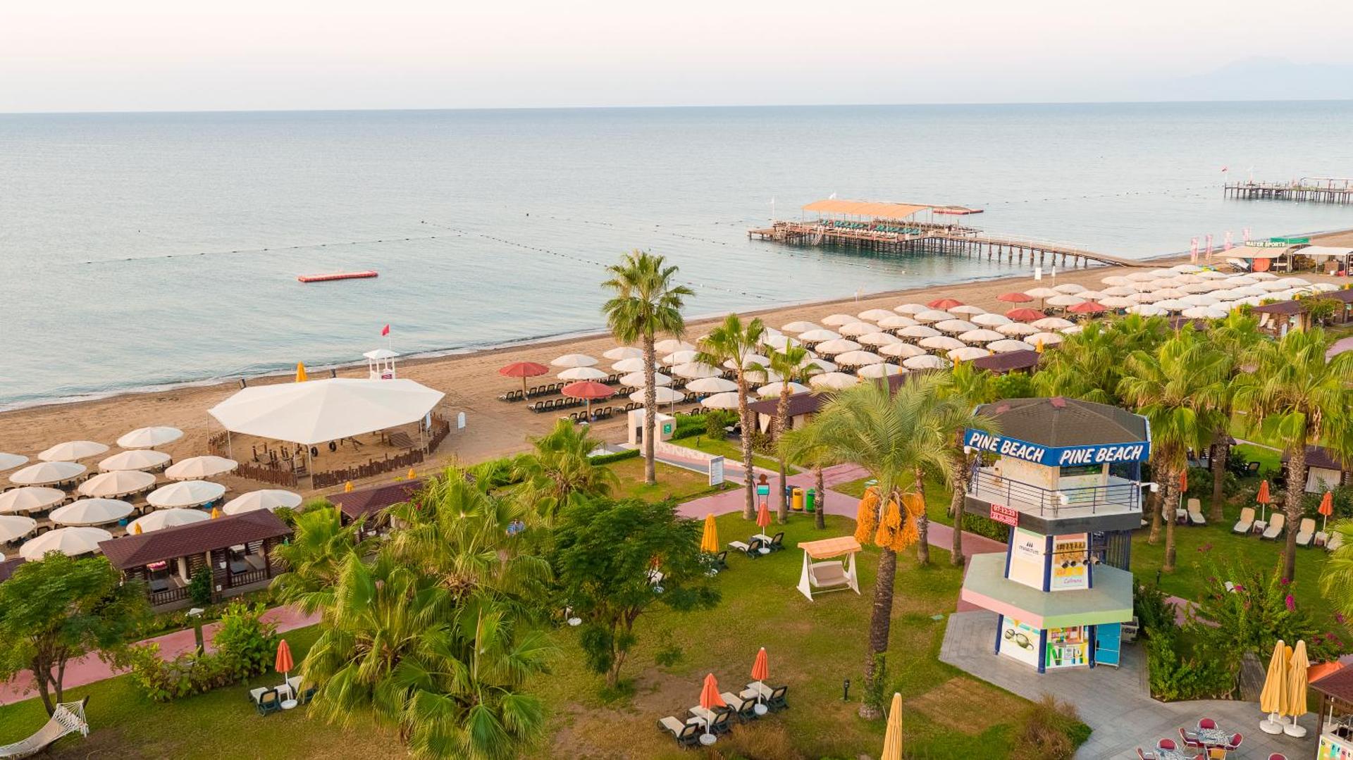 Pine Beach Belek viesnīcas dārza un pludmales zona ar palmām, bāru un koka piestātni Vidusjūras piekrastē