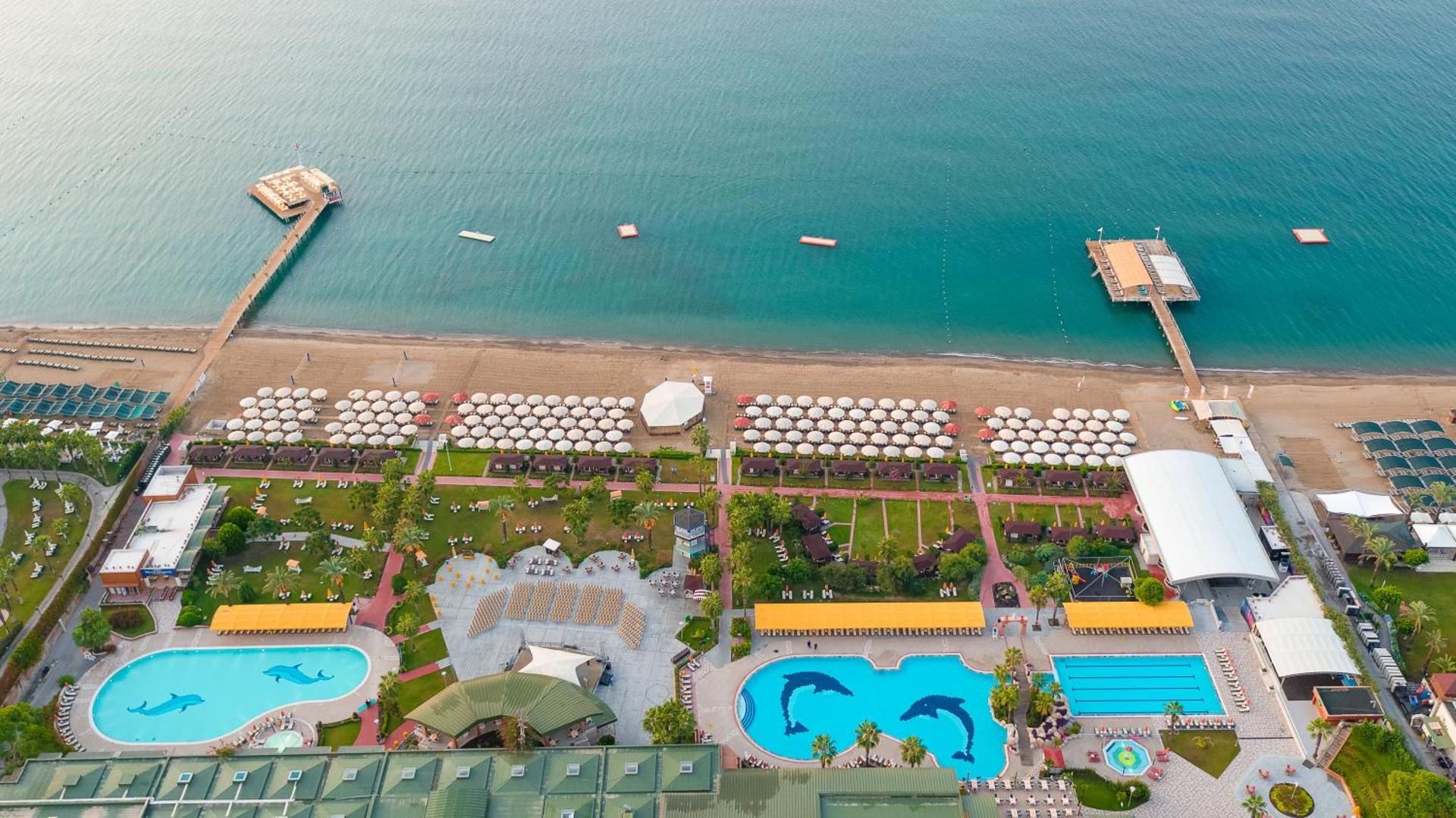 Skats no augšas uz Pine Beach Belek pludmali ar saulessargiem, divām koka piestātnēm un vairākiem āra baseiniem piekrastē.