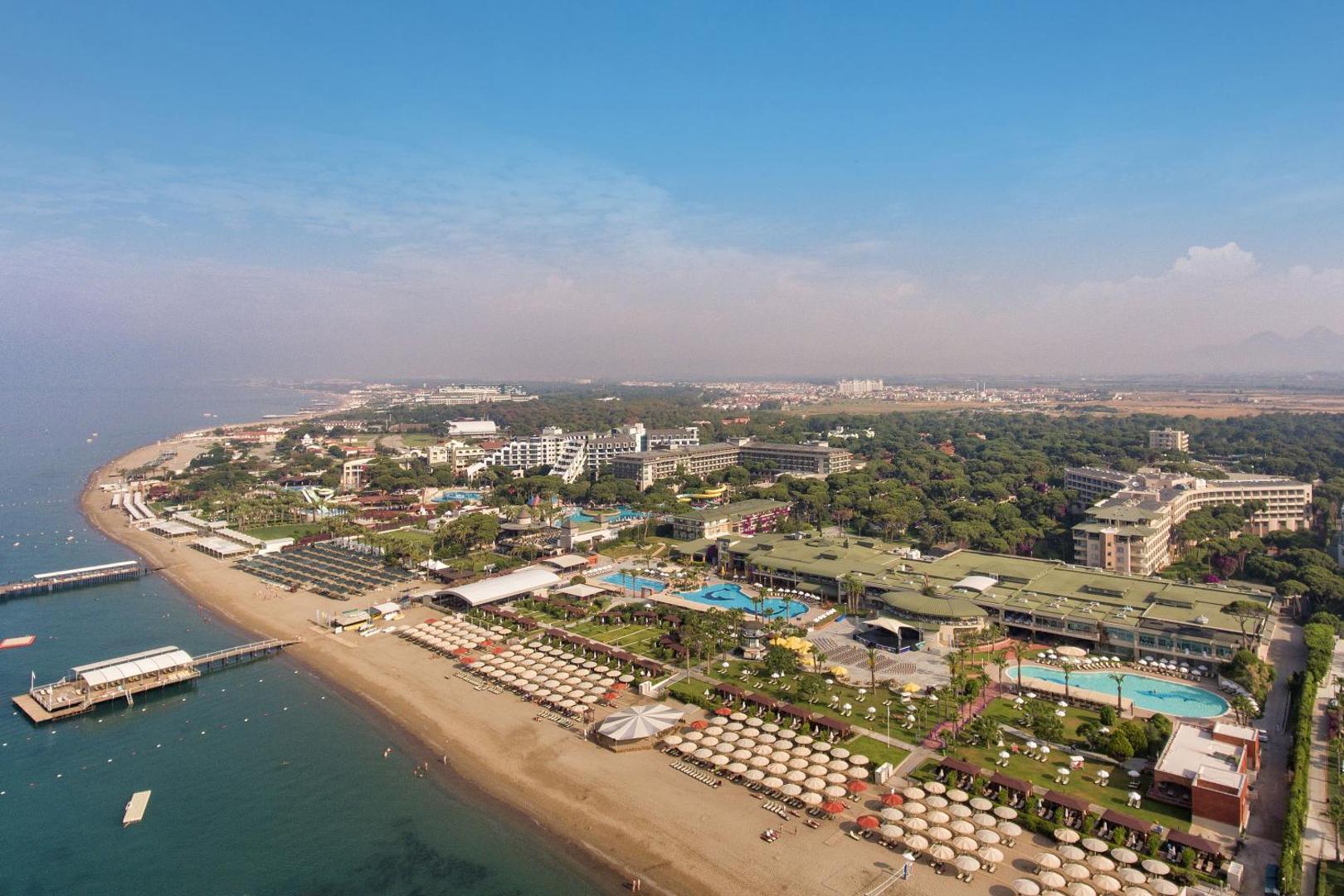 Aerofoto skats uz Pine Beach Belek kūrortu ar plašo smilšu pludmali, piestātnēm un zaļo priežu meža teritoriju Belekā.