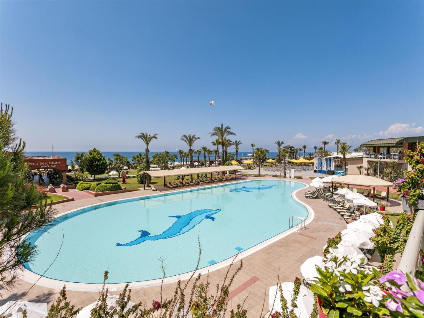 Skats no augšas uz Pine Beach Belek baseinu ar delfīnu mozaīku, saulessargiem un jūras piekrasti tālumā