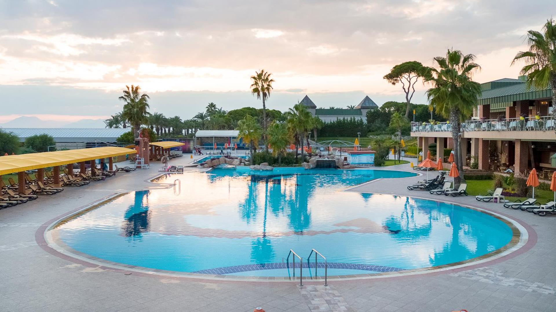 Pine Beach Belek atpūtas zona ar lielu baseinu, dzeltenām nojumēm un zaļojošu dārzu saulainā pēcpusdienā