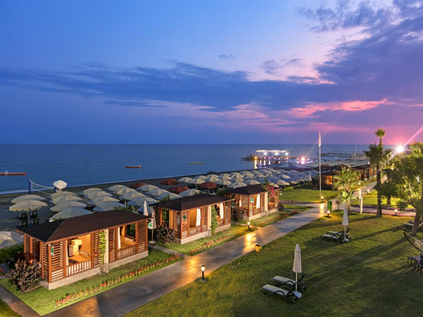 Pine Beach Belek dārza zona saulrietā ar apgaismotiem koka bungalo, zālāju un skatu uz jūru Belekas reģionā