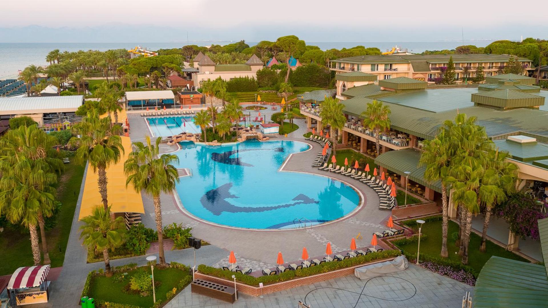 Pine Beach Belek viesnīcas centrālais āra baseins ar delfīnu mozaīku, ko ieskauj palmas un sauļošanās krēsli Antālijas reģionā, Turcijā.
