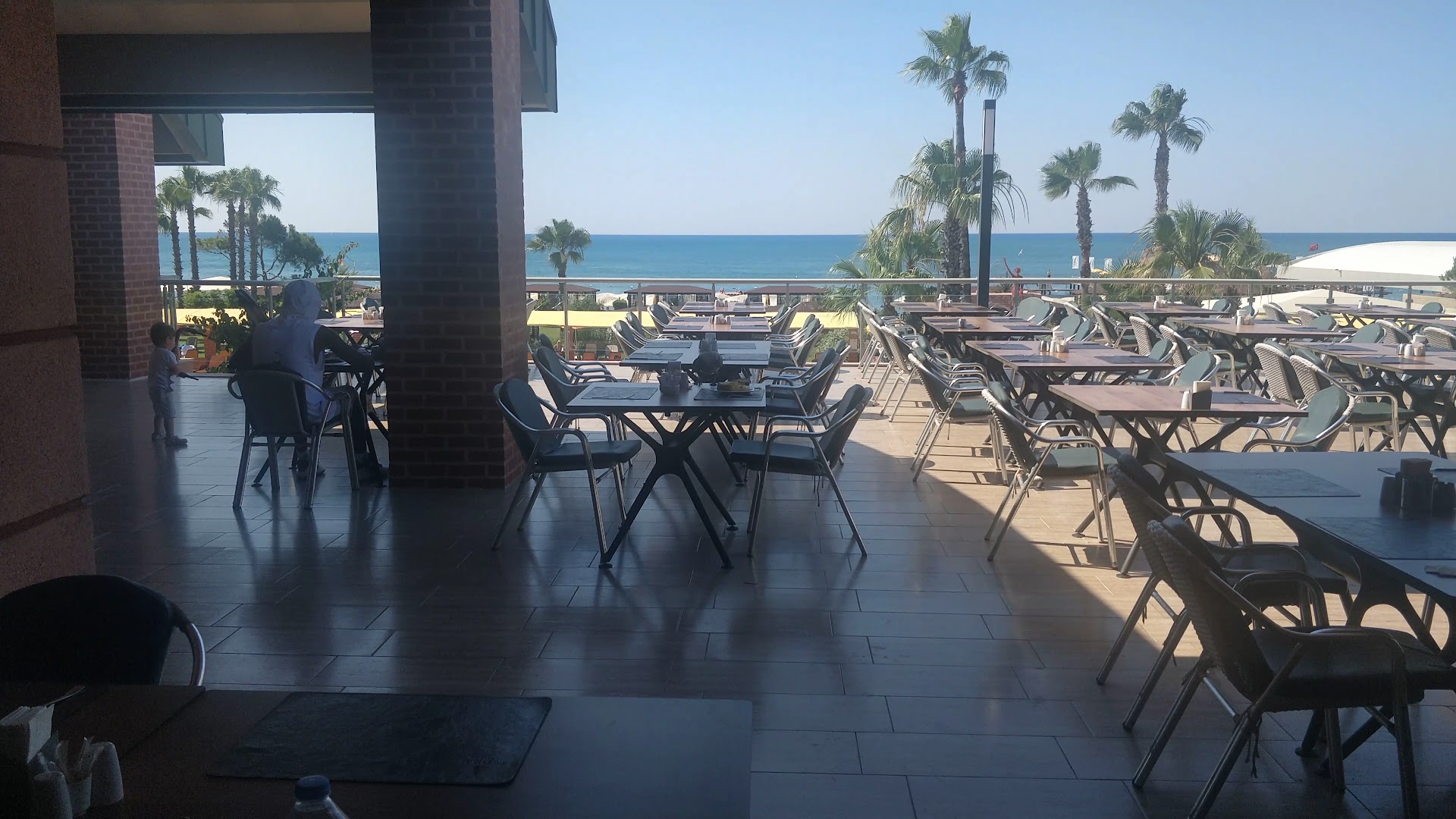 Pine Beach Belek āra terase ar galdiņiem un skatu uz Vidusjūru un palmām saulainā dienā