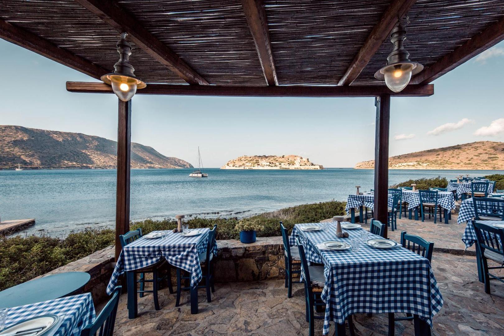Phaea Blue restorāna āra terase ar tradicionāliem rūtainiem galdautiem un panorāmas skatu uz Spinalongas salu