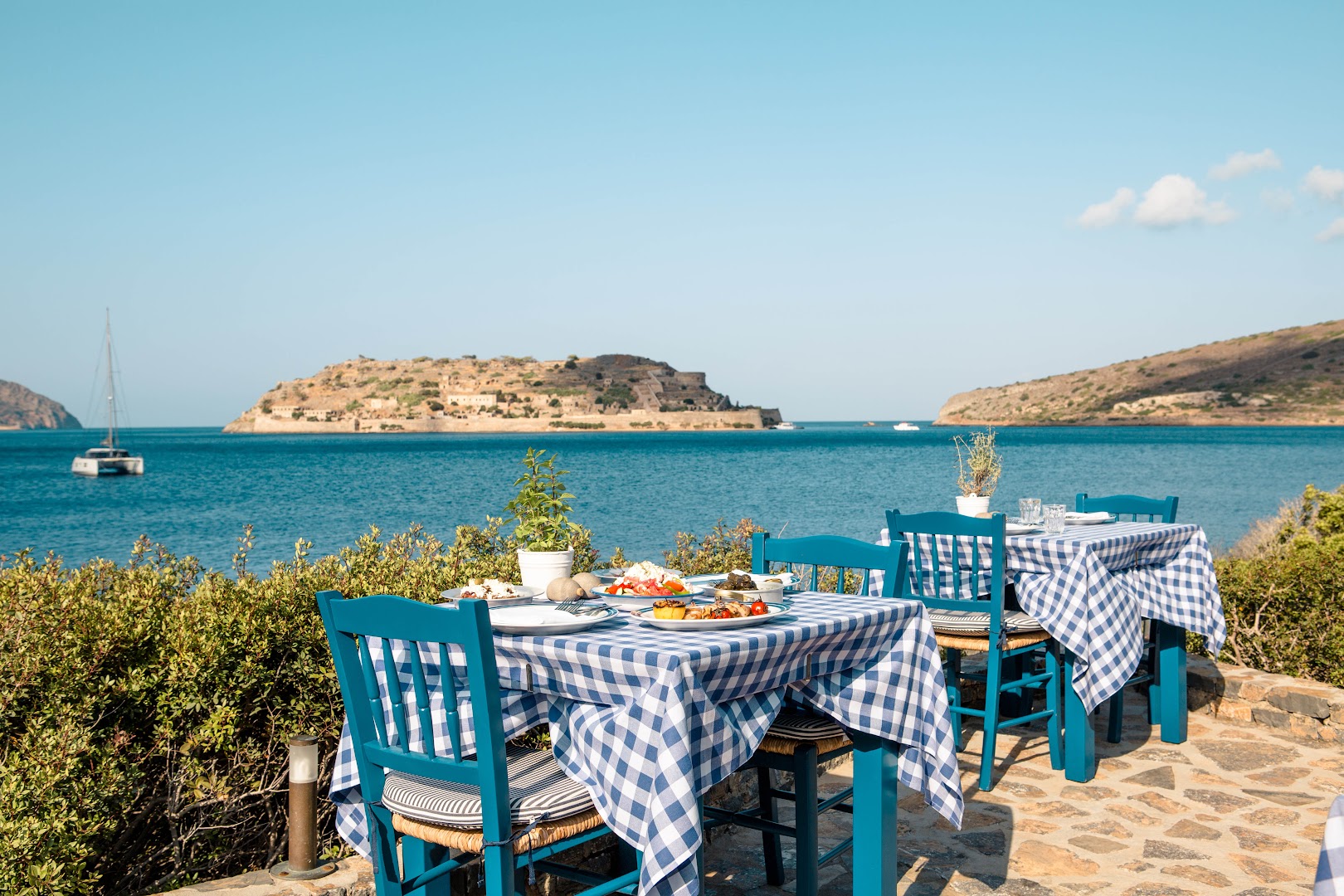 Tradicionāla grieķu stila āra kafejnīca Phaea Blue piekrastē ar ziliem koka krēsliem un skatu uz Spinalongas salu un līci