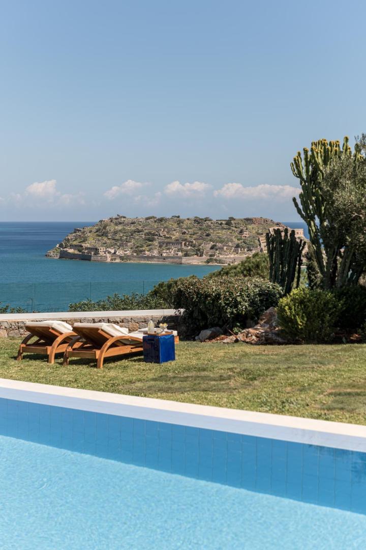 Sauļošanās krēsli uz zālāja pie Phaea Blue baseina ar tiešu skatu uz Spinalongas vēsturisko salu un jūru
