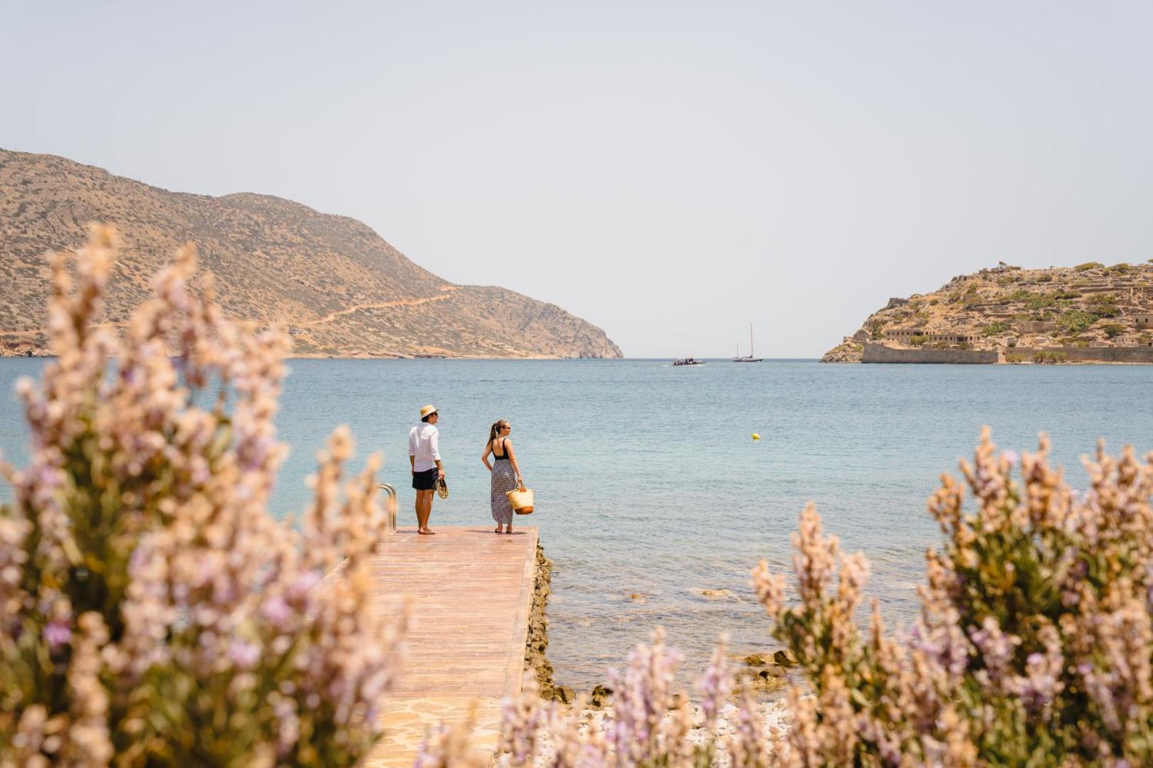 Pāris stāv uz koka piestātnes Phaea Blue kūrortā, raugoties uz jūras līci un Spinalongas salu caur ziedošiem krūmiem