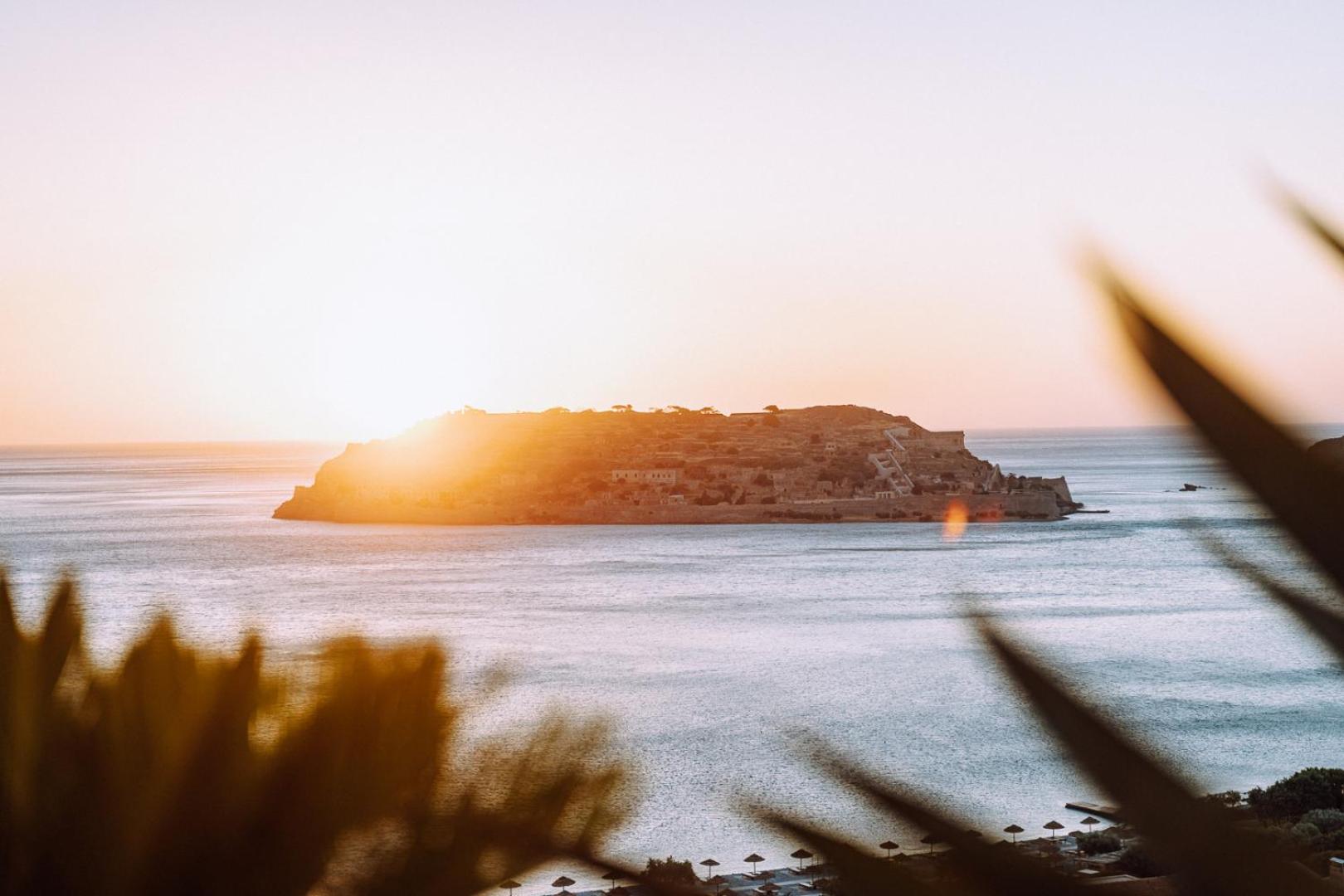 Saulrieta gaisma pār Spinalongas salu un jūru, skatoties no Phaea Blue kūrorta piekrastes Krētā