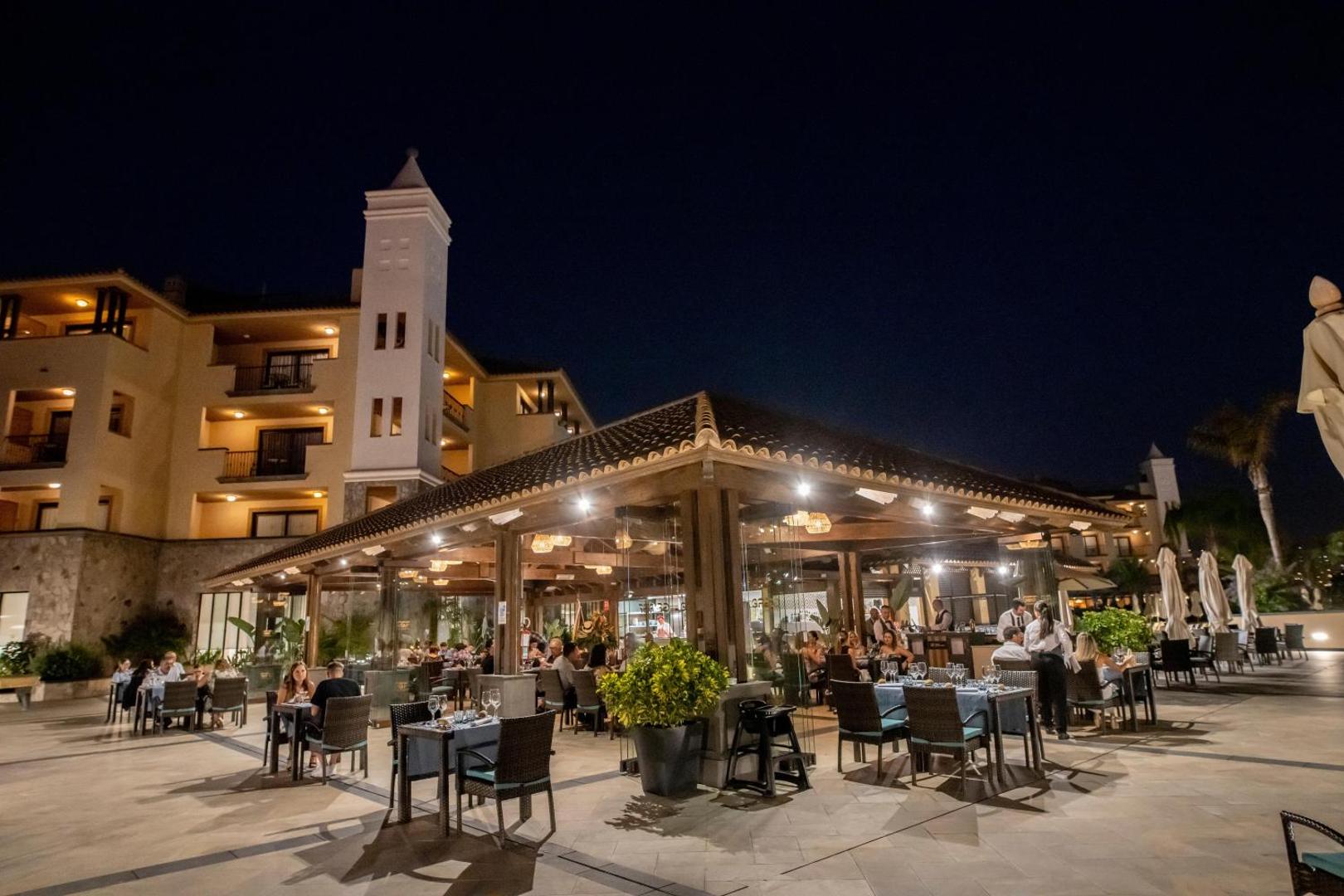 GF Gran Costa Adeje āra restorāna terase vakarā ar galdiņiem un viesiem pie viesnīcas ēkas Costa Adeje kūrortā