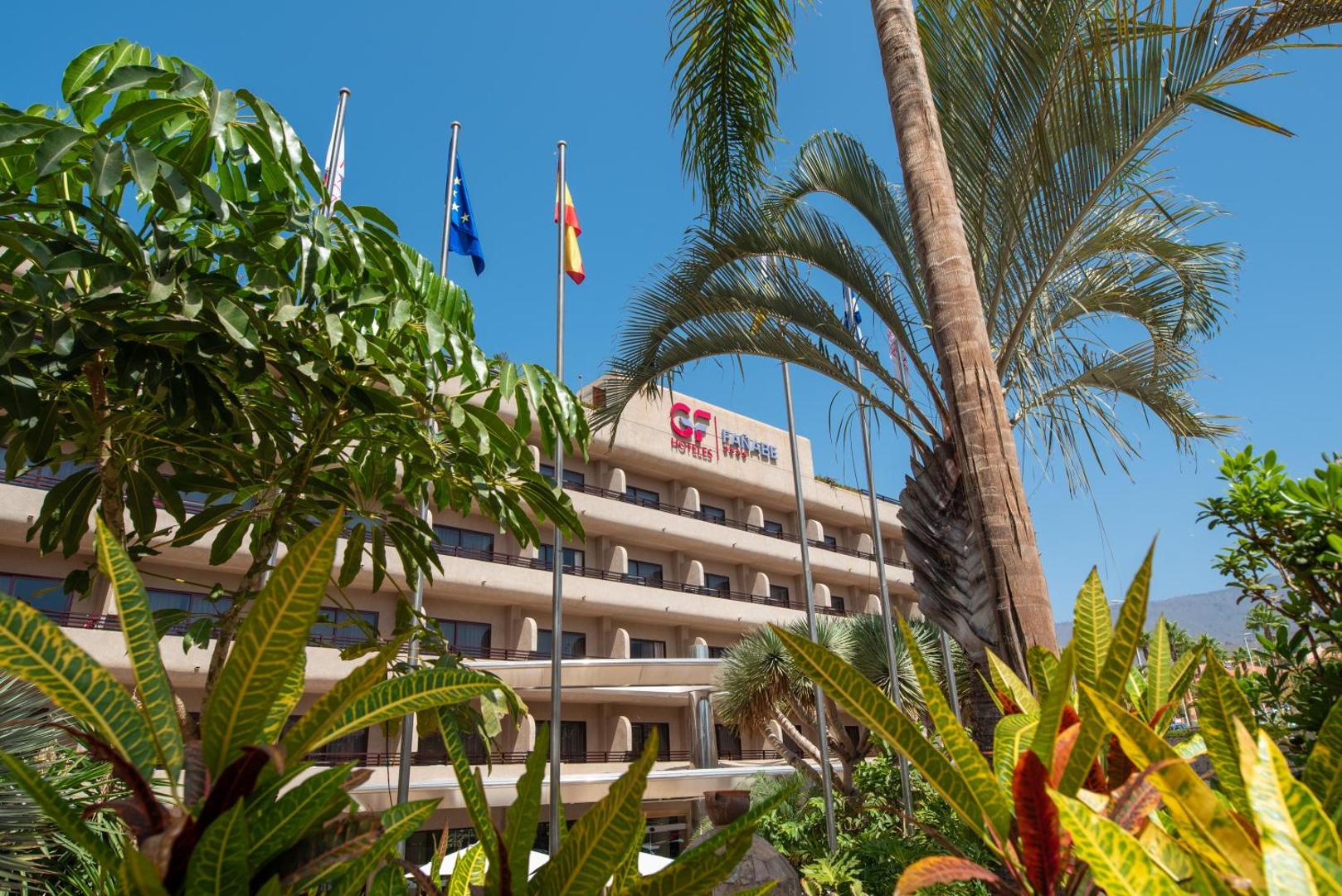 GF Fañabé ieeja ar palmām un karogiem Tenerifes piekrastē — skats uz viesnīcas korpusu caur tropu zaļumiem
