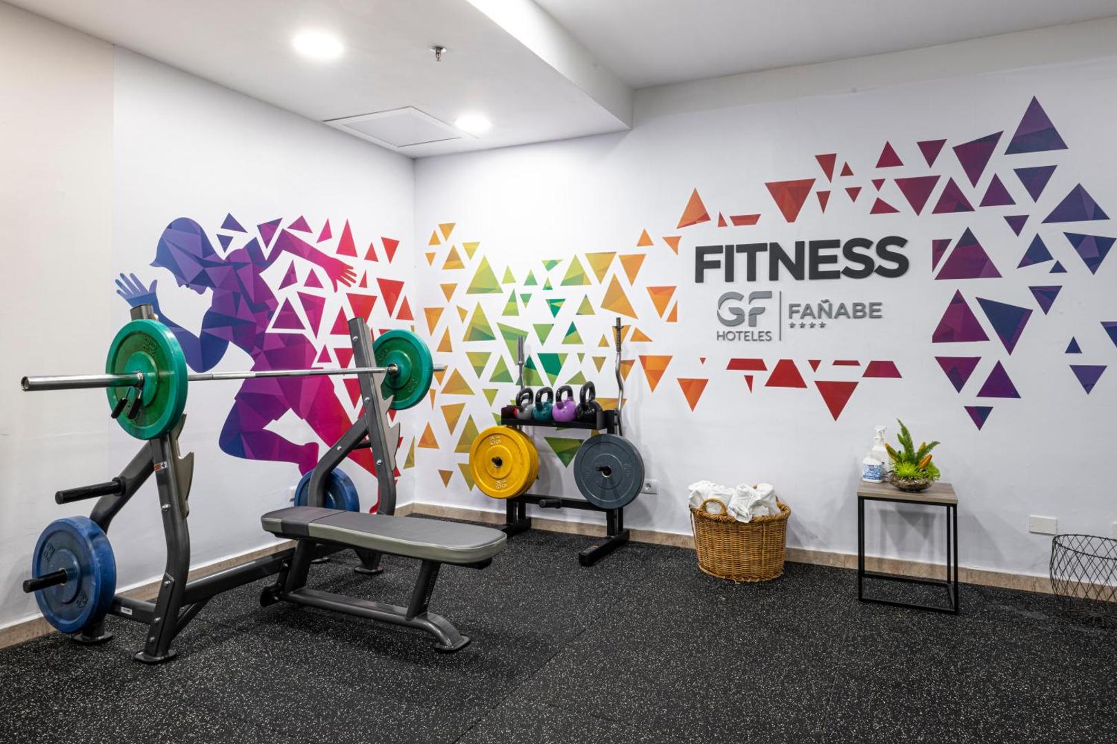 GF Fañabe viesnīcas fitness centrs Tenerifē ar svaru stieni, soliņu un krāsainiem svaru diskiem pie sienas ar grafiti