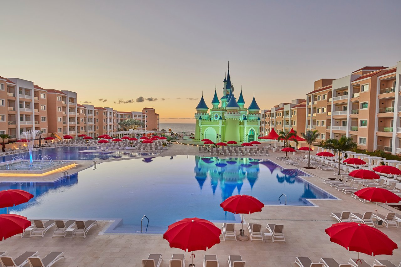Bahia Principe Fantasia Tenerife plašs āra baseins ar strūklaku, sarkanajiem saulessargiem un centrālo pasaku pili krēslas stundā.