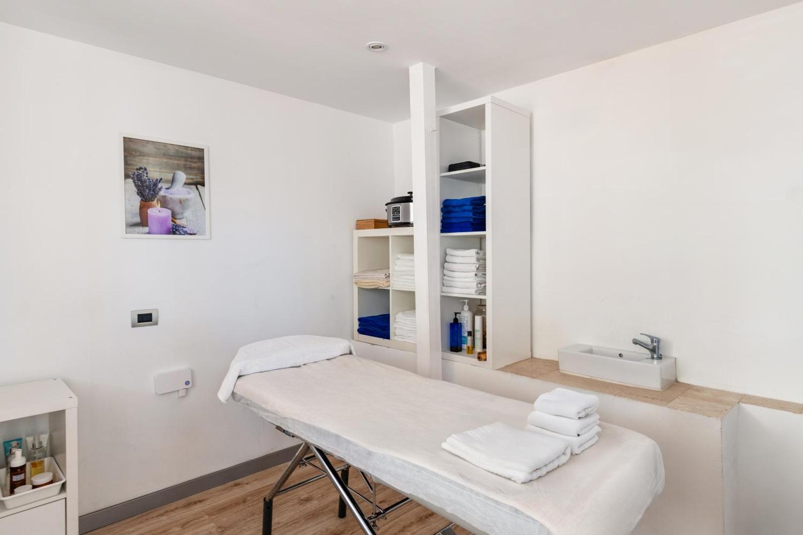Viesnīcas Catalonia Oro Negro spa procedūru telpa ar masāžas galdu, baltiem dvieļiem un relaksējošu atmosfēru mierpilnai atpūtai