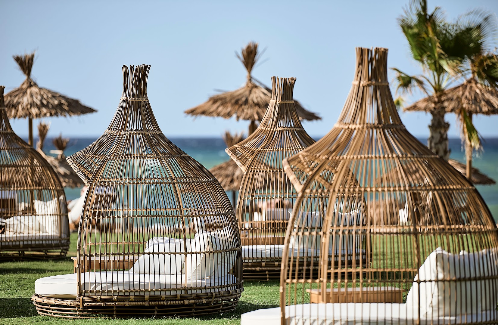 Pepper Sea Club dizaina atpūtas krēsli bungalo stilā ar baltiem spilveniem zālājā pie jūras — saulessargi un palmas fonā