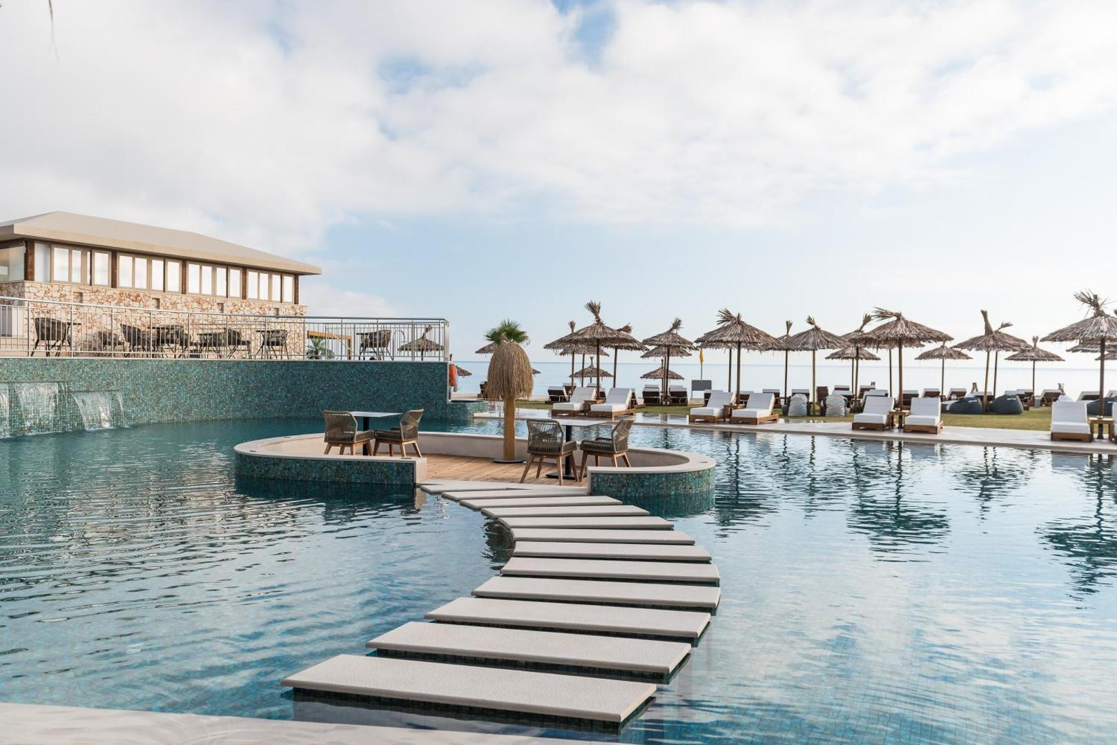 Pepper Sea Club dizaina baseins ar akmens taku un atpūtas zonu — fonā pludmales saulessargi un jūra Krētas piekrastē