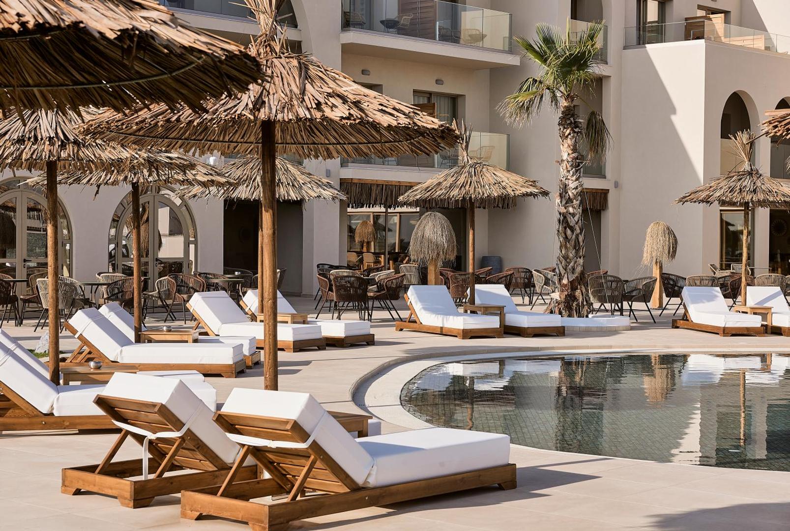Pepper Sea Club Hotel āra baseins ar koka sauļošanās krēsliem un salmu saulessargiem Krētā, Grieķijā — moderna atpūtas zona pie viesnīcas korpusa