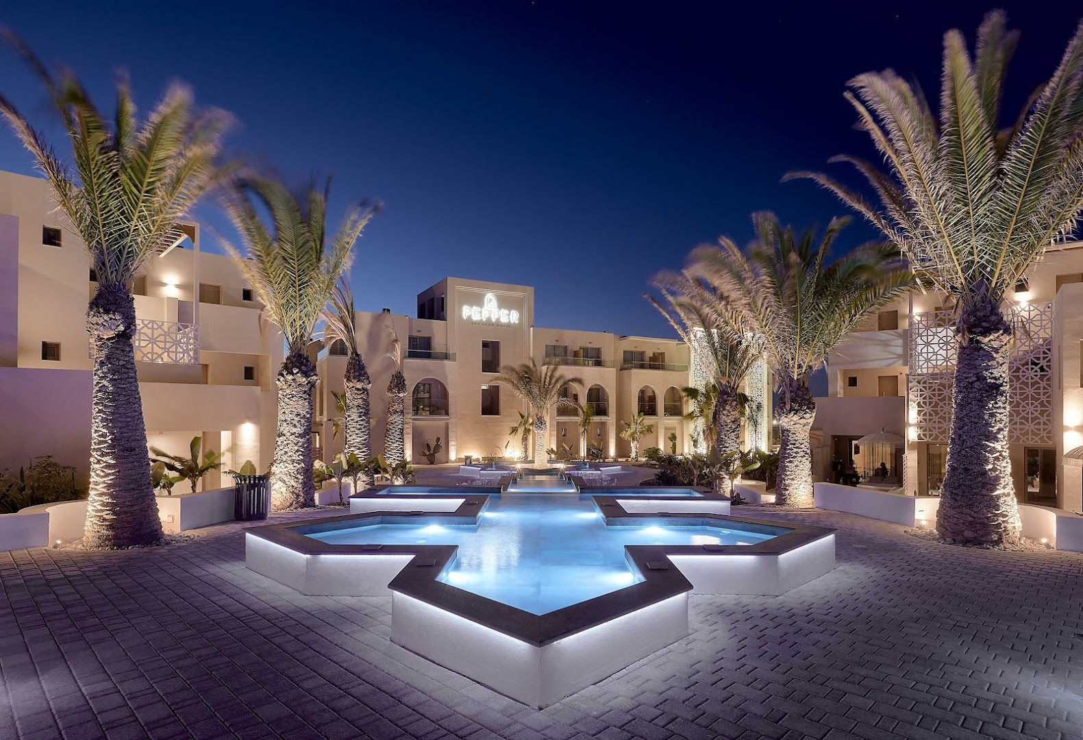 Pepper Sea Club Hotel centrālais pagalms vakarā ar izgaismotu dekoratīvu baseinu, palmām un viesnīcas fasādi Krētā