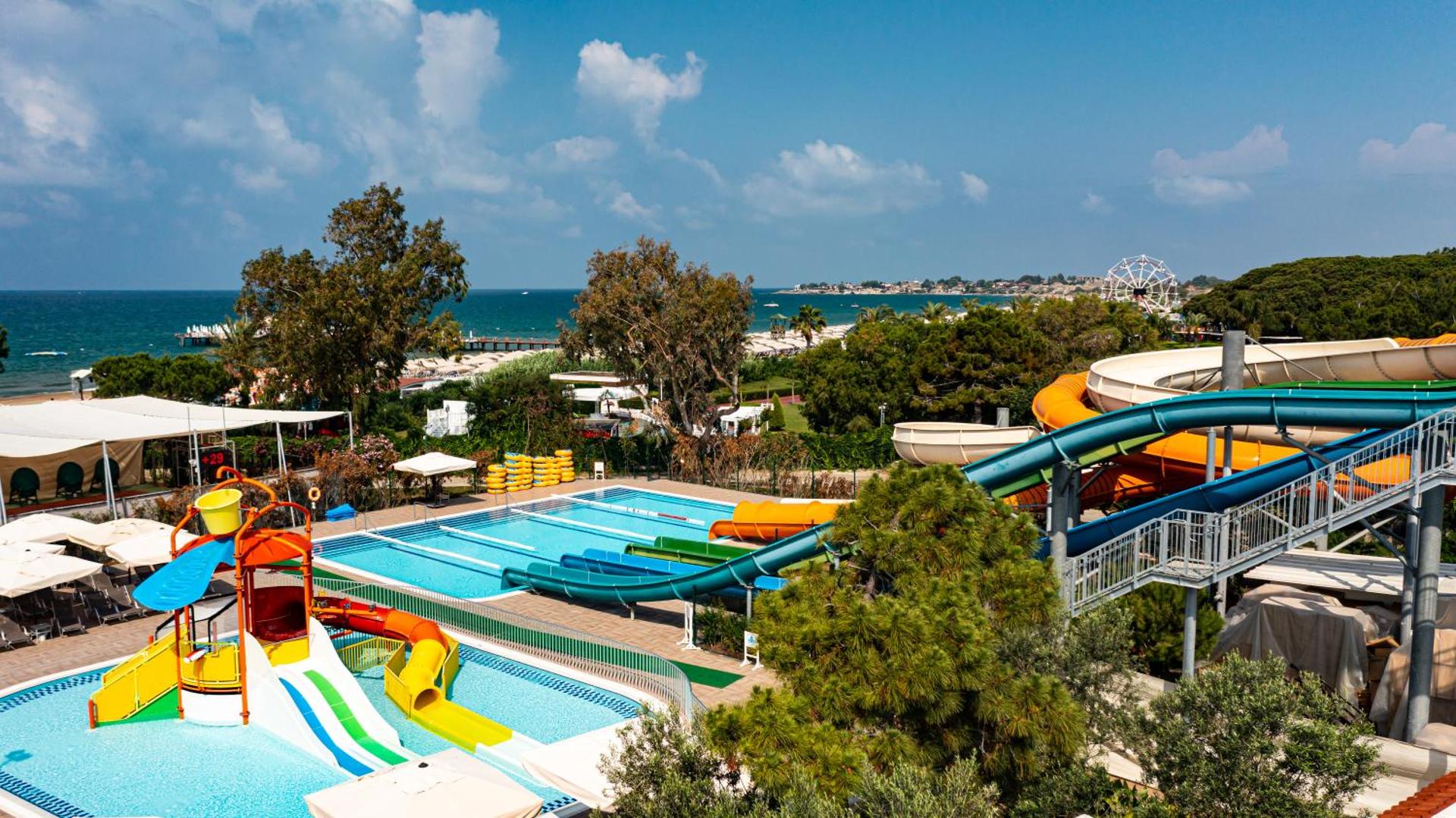 Ali Bay Sorgun viesnīcas akvaparks ar krāsainiem ūdens slidkalniņiem un baseiniem piekrastē, fonā redzama jūra un panorāmas rats.