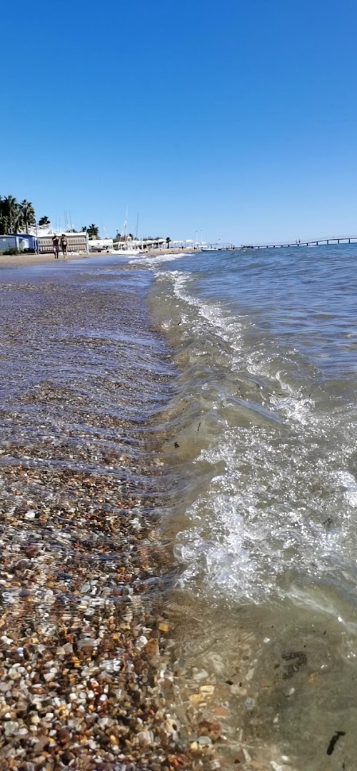 Tuvplāns ar jūras viļņiem Ali Bay Resort Sorgun oļu un smilšu pludmalē saulainā dienā ar skatu uz piestātni tālumā.