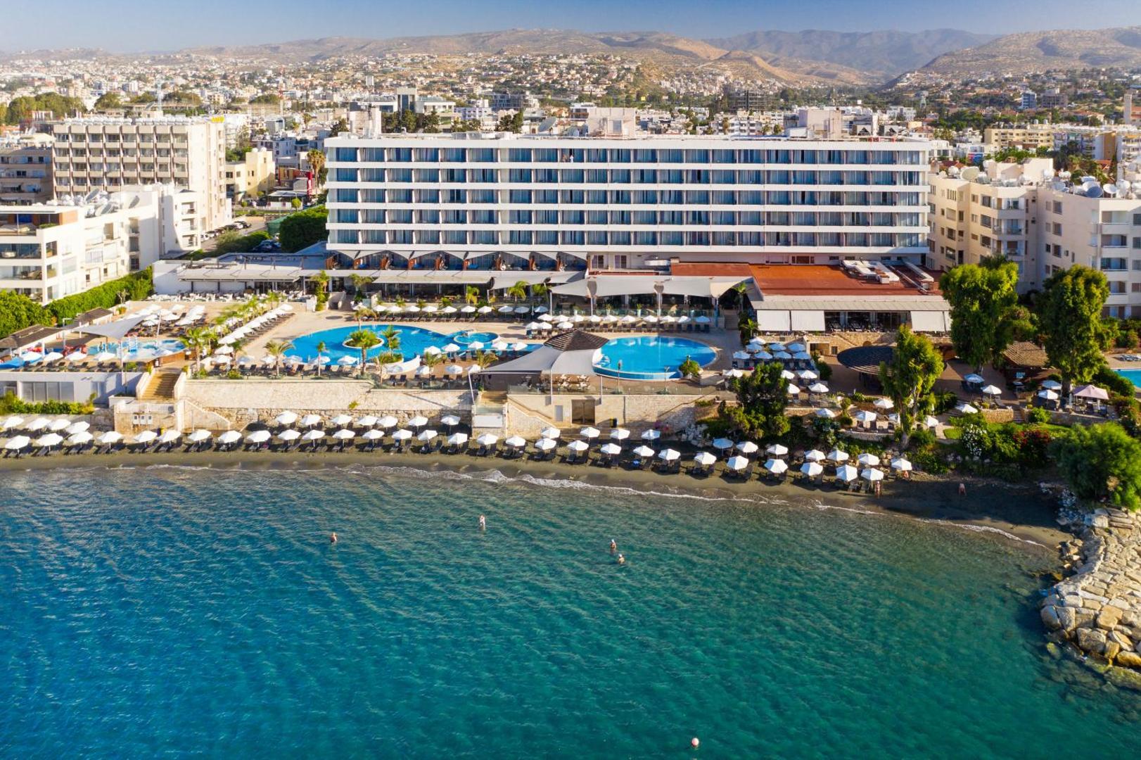 The Royal Apollonia panorāmas skats uz galveno ēku, baseiniem un privāto pludmali Limasolā — plaša atpūtas zona ar saulessargiem