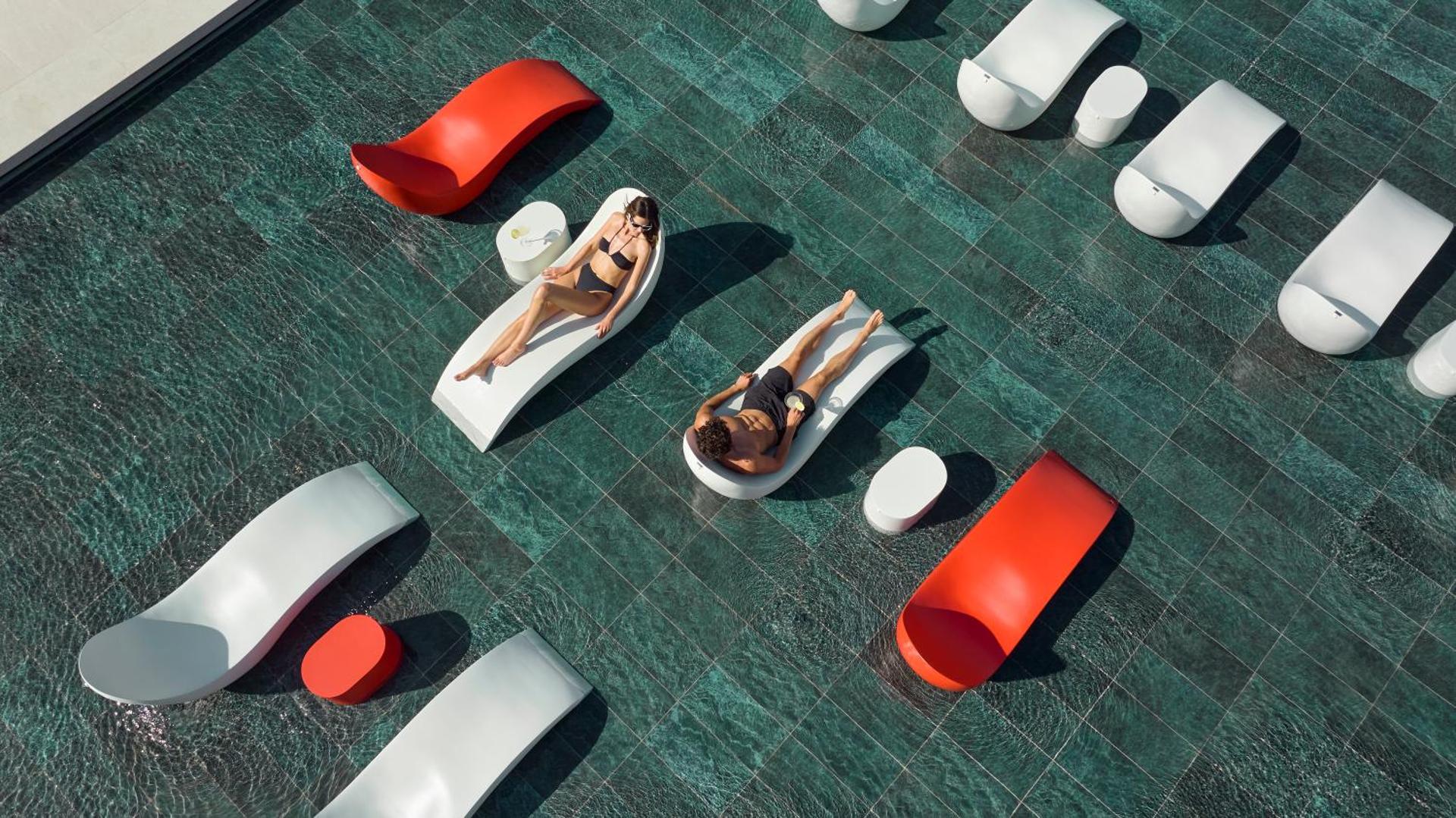 Anda Barut Collection moderni dizaina sauļošanās krēsli baseinā — skats no augšas uz pāri, kas atpūšas uz baltiem un sarkaniem zviļņiem