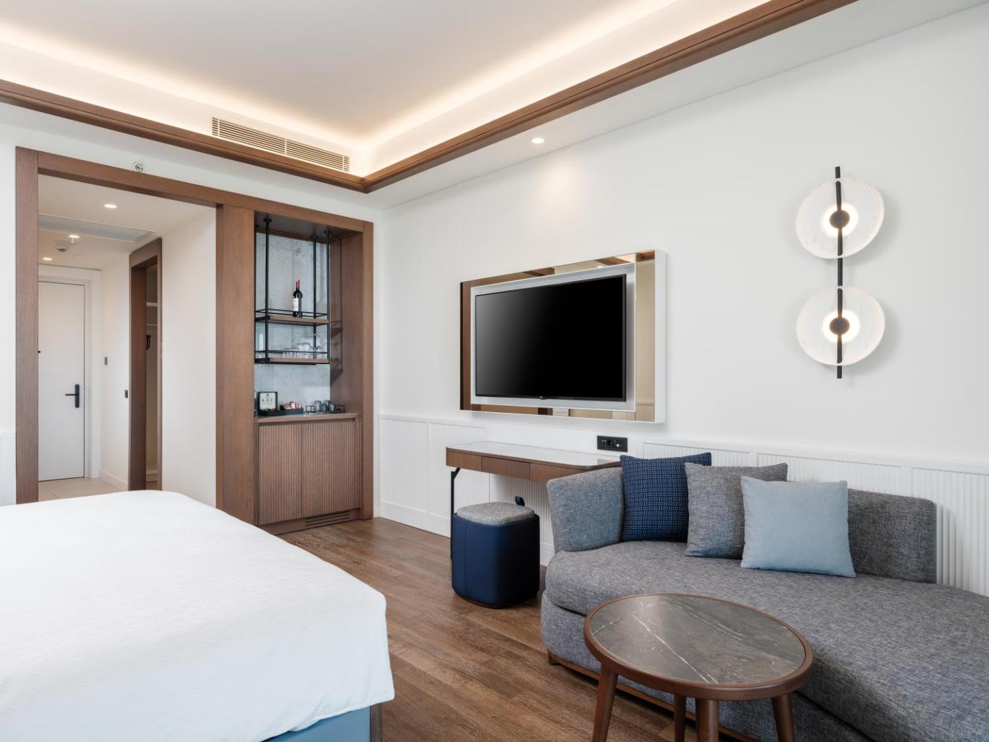 Anda Barut Collection moderns viesnīcas numurs ar gaišu interjeru, pelēku dīvānu, televizoru un elegantiem koka apdares elementiem