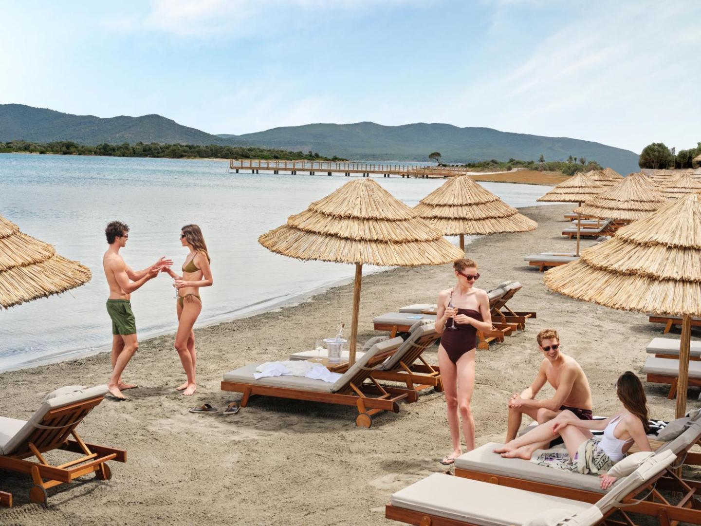Viesi atpūšas Anda Barut Collection pludmalē pie lēzenas ieejas jūrā — koka sauļošanās krēsli un dabiski salmu saulessargi