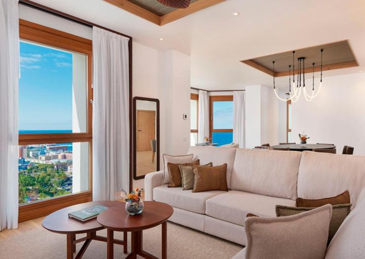 Gran Hotel Taoro luksusa numura viesistaba ar gaišu dīvānu, koka galdiņiem un plašiem logiem ar skatu uz okeānu Tenerifē