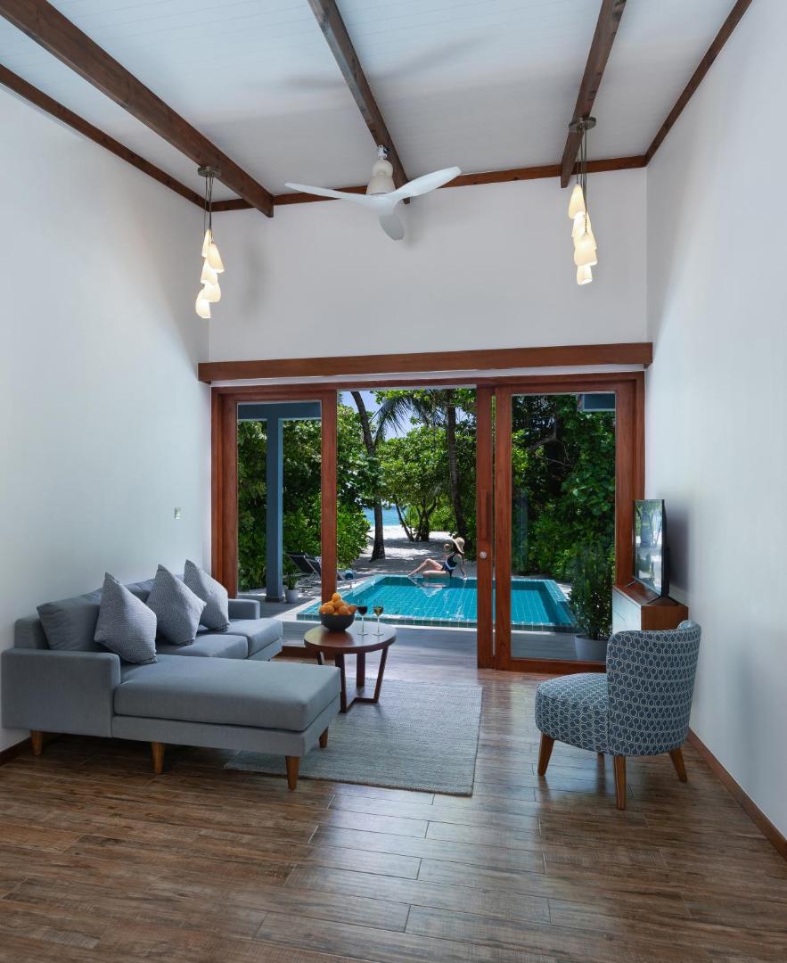 Ifuru Island Maldives villas dzīvojamā zona ar pelēku dīvānu un skatu uz privāto baseinu caur stikla durvīm