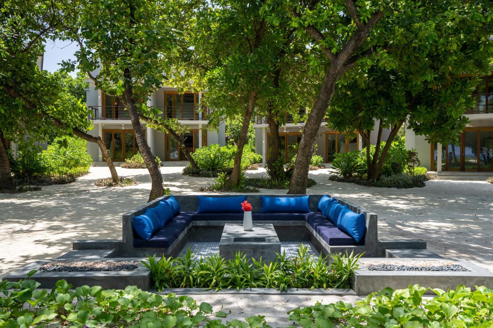 Ifuru Island Maldives atpūtas zona dārzā ar mīkstiem dīvāniem un ziliem spilveniem, ko ieskauj zaļojoši tropu koki