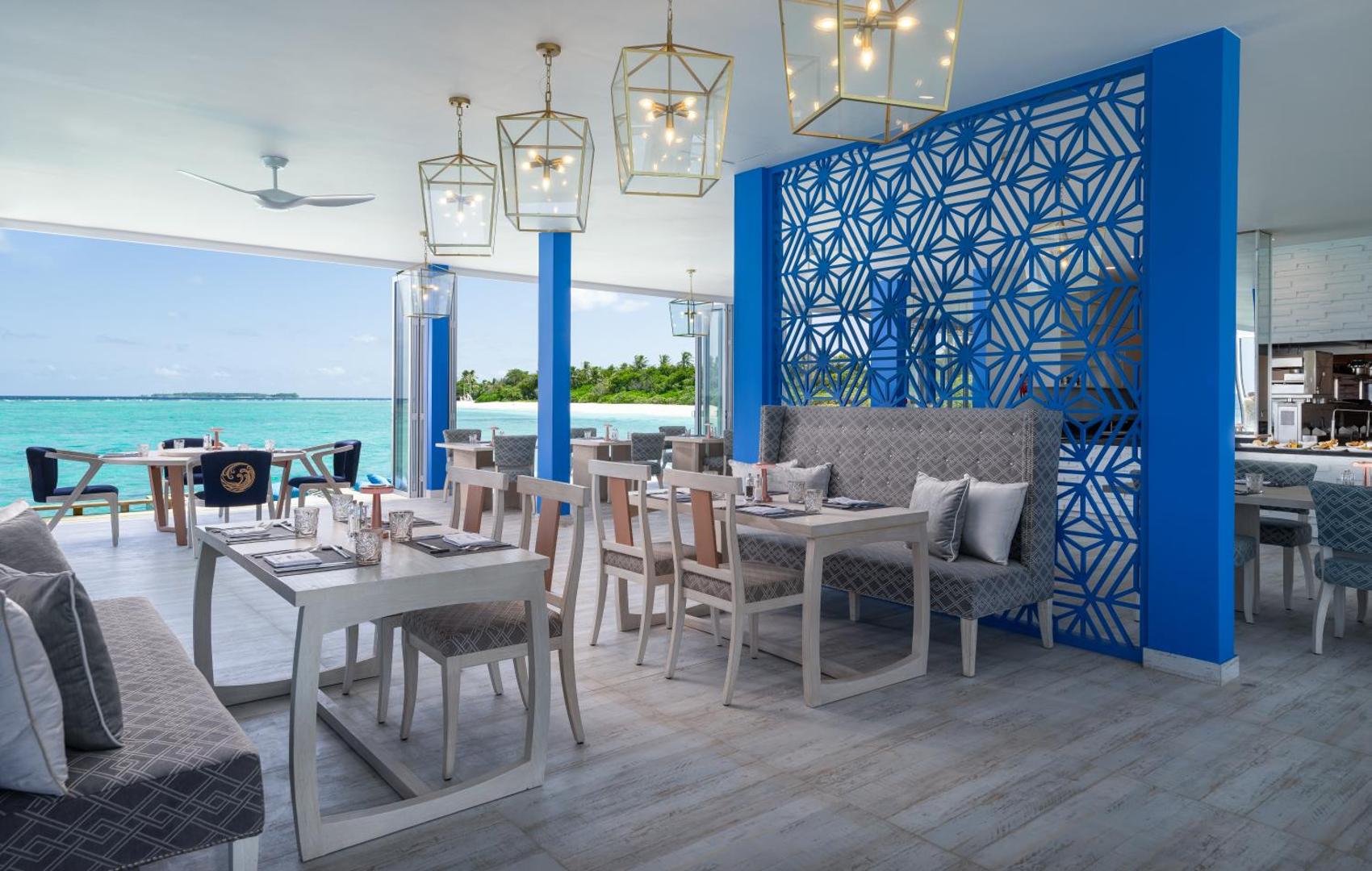 Ifuru Island Maldives moderns restorāns ar gaišām mēbelēm, ziliem akcentiem un skatu uz tirkīzzilo okeānu