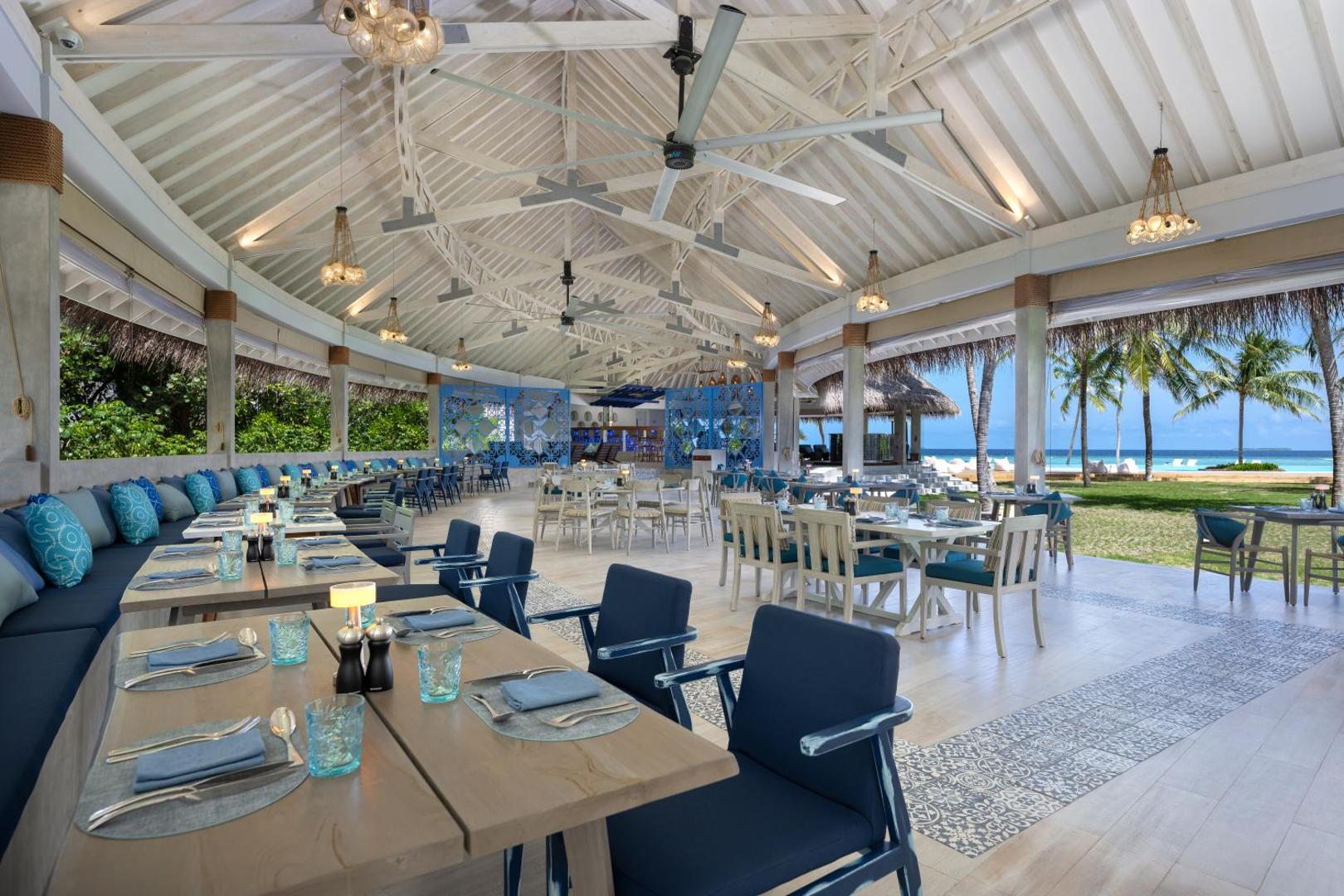 Ifuru Island Maldives plaša restorāna terase ar zilām mēbelēm un skatu uz pludmali — atvērta tipa arhitektūra un palmas