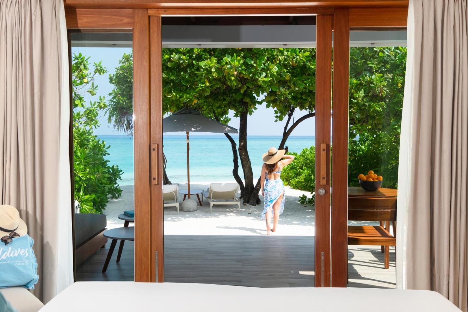 Skats no Ifuru Island Maldives numura uz balto smilšu pludmali — sieviete cepurē dodas uz okeāna krastu caur zaļojošu dārzu