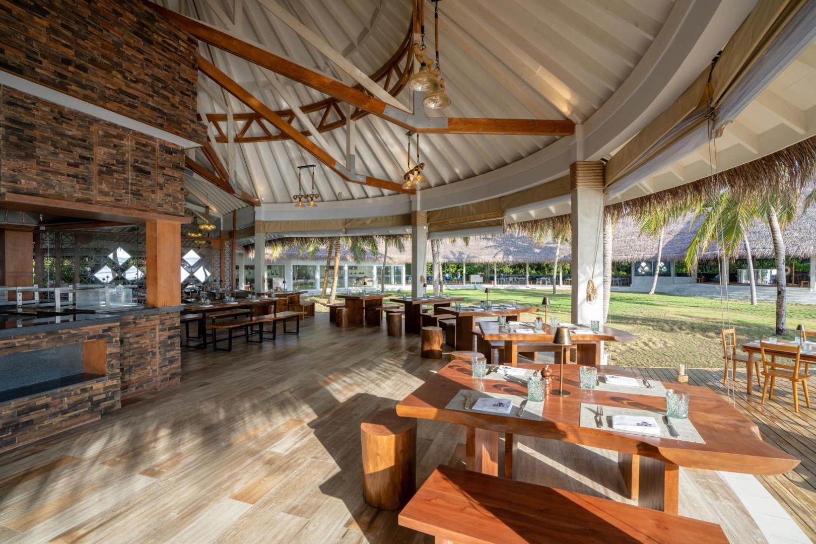Ifuru Island Maldives restorāns ar koka mēbelēm un akmens apdari zem augstiem griestiem — skats uz zaļo zonu un palmām