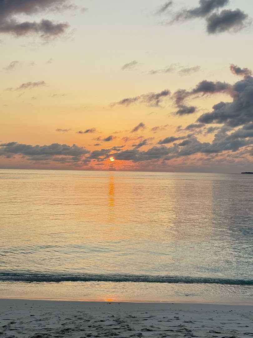 Saulriets Ifuru Island Maldives kūrorta pludmalē ar zeltainām debesīm, mākoņiem un mierīgu okeāna ūdeni piekrastē