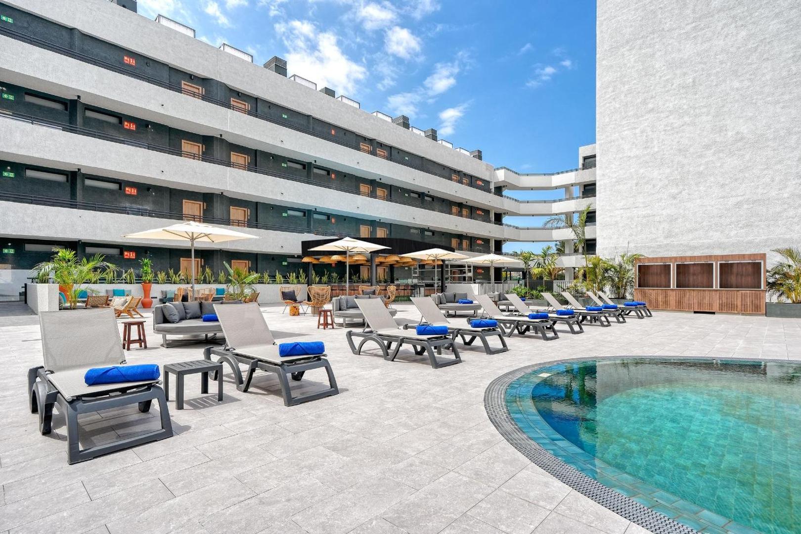 Hovima Suites Costa Adeje sauļošanās terase pie baseina ar pelēkiem zviļņiem un ziliem dvieļiem, skats uz viesnīcas korpusu un bāru