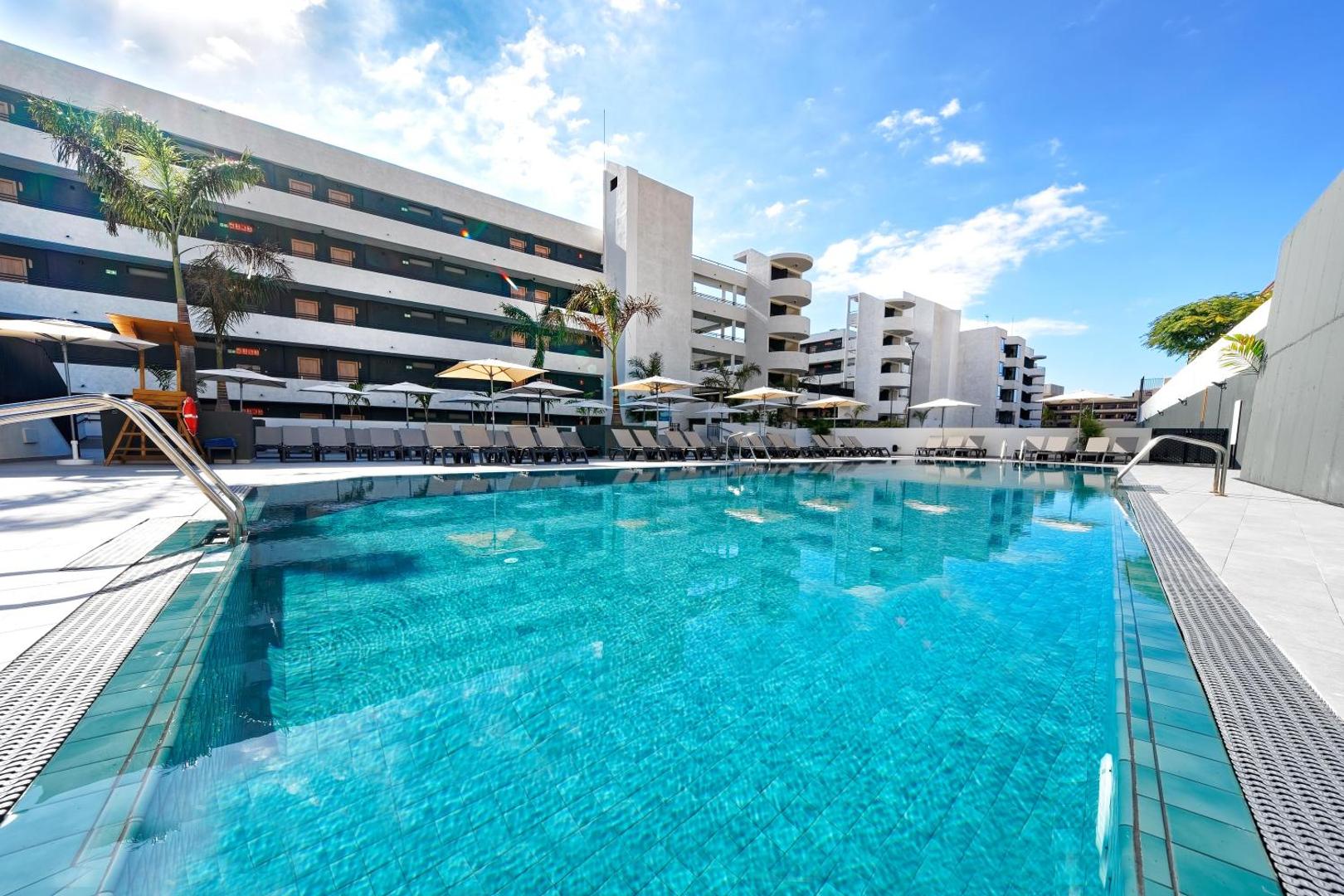 Hovima Suites Costa Adeje āra baseins ar dzidru ūdeni un skatu uz viesnīcas ēku zem skaidrām debesīm Tenerifē