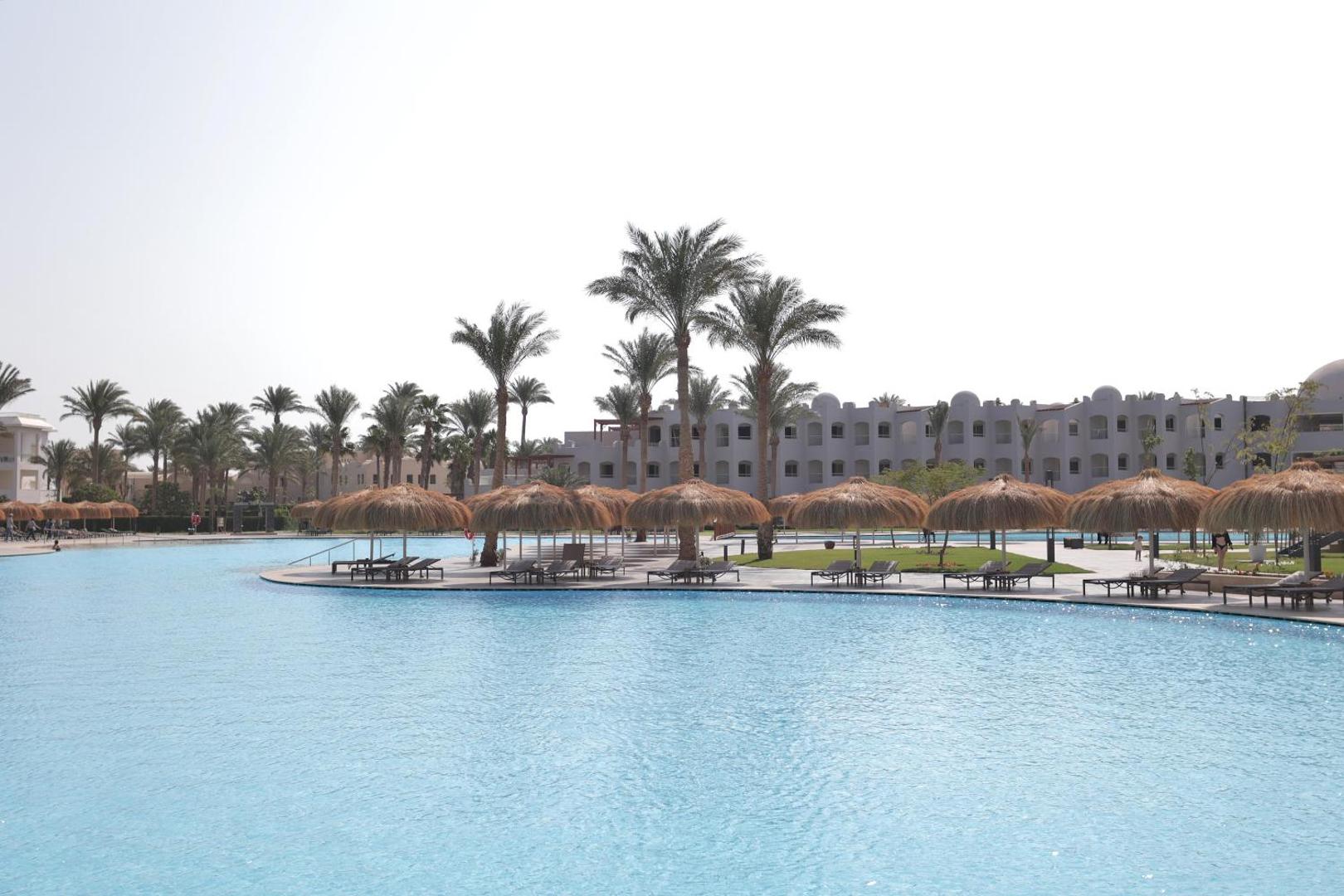 Skats uz Xanadu Club Makadi Bay baseinu un viesnīcas ēkām caur palmu rindām un saulessargiem Makadibejā