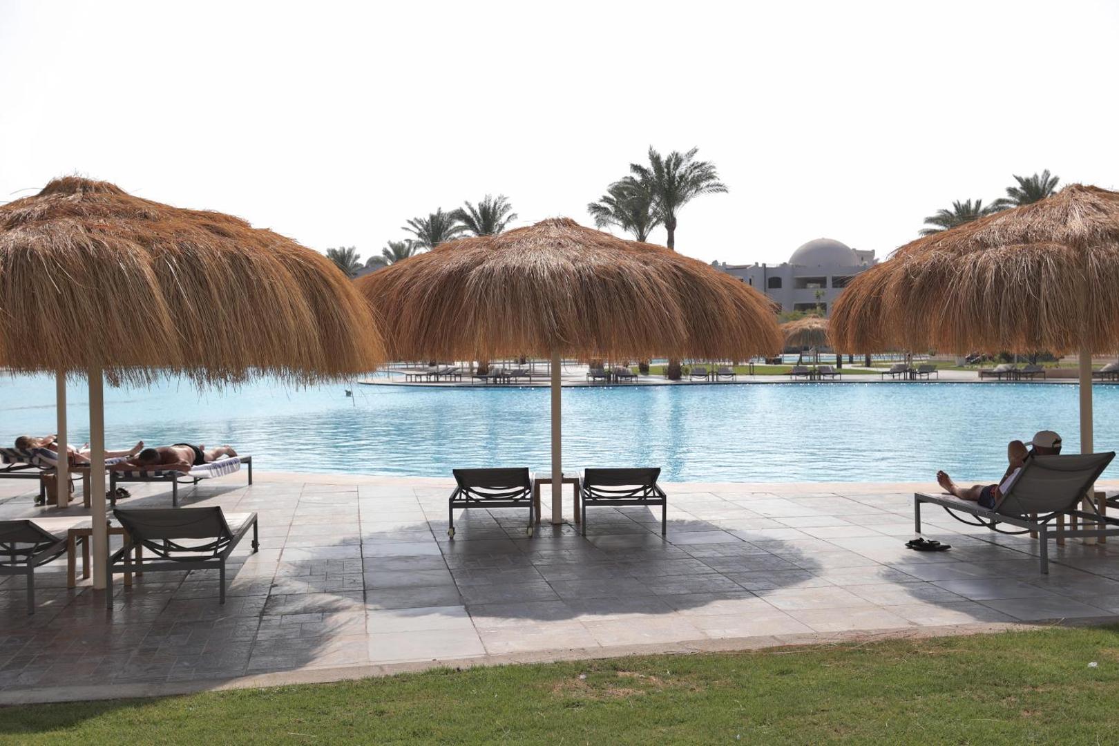 Atpūtas krēsli zem salmu saulessargiem pie Xanadu Club Makadi Bay baseina ar skatu uz kūrorta teritoriju