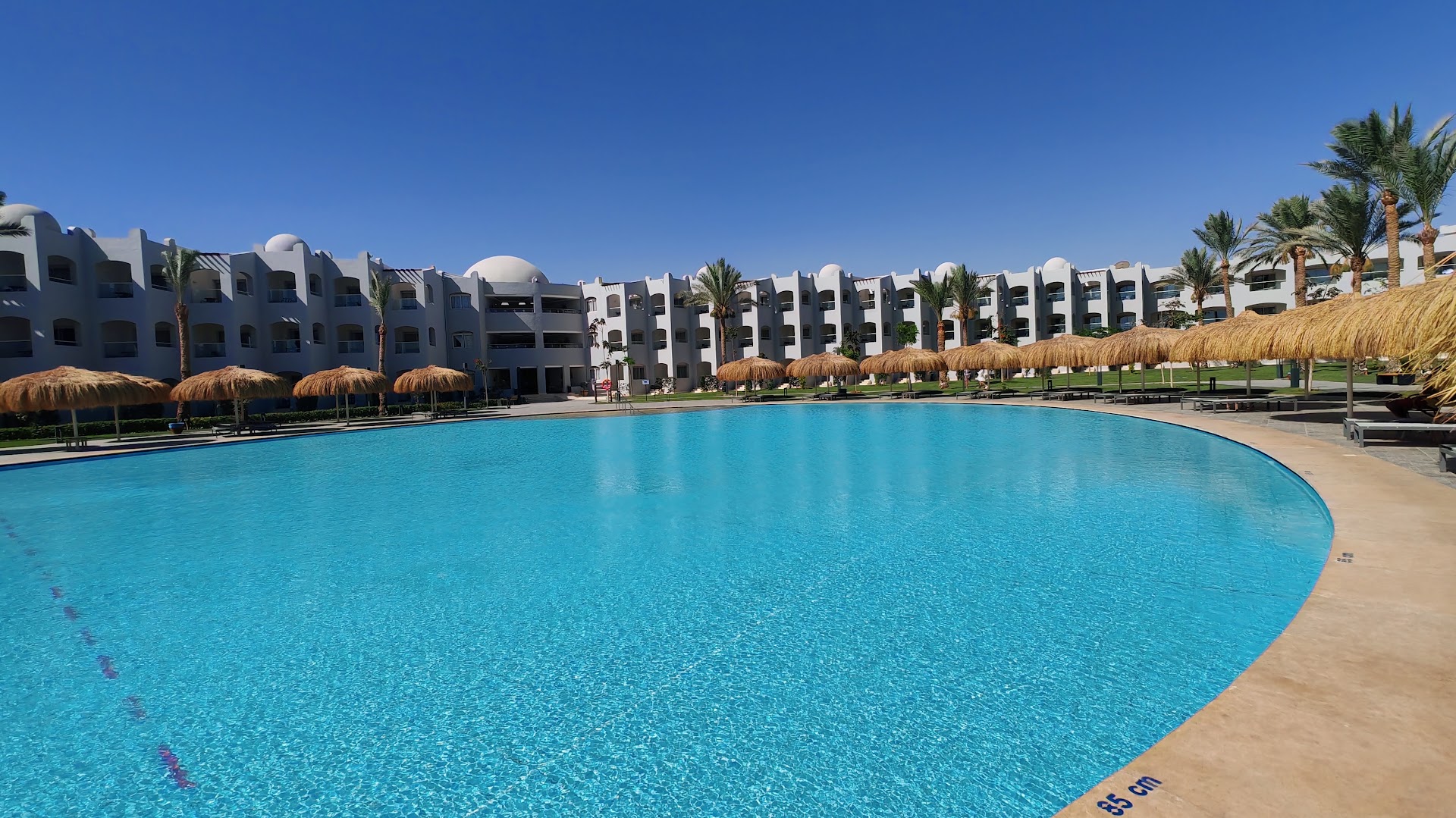 Xanadu Club Makadi Bay galvenais baseins ar dzidru ūdeni, salmu saulessargiem un viesnīcas korpusiem fonā saulainā dienā