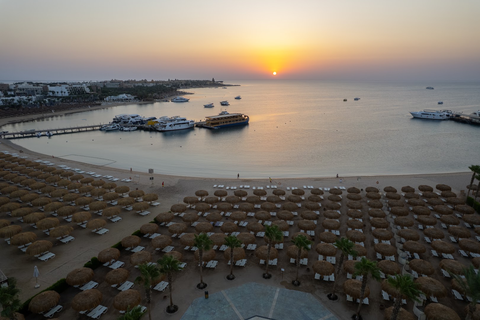 Xanadu Club Makadi Bay panorāmas skats uz pludmali saulrieta laikā Hurgadā — simtiem saulessargu, jahtu piestātne un Sarkanā jūra
