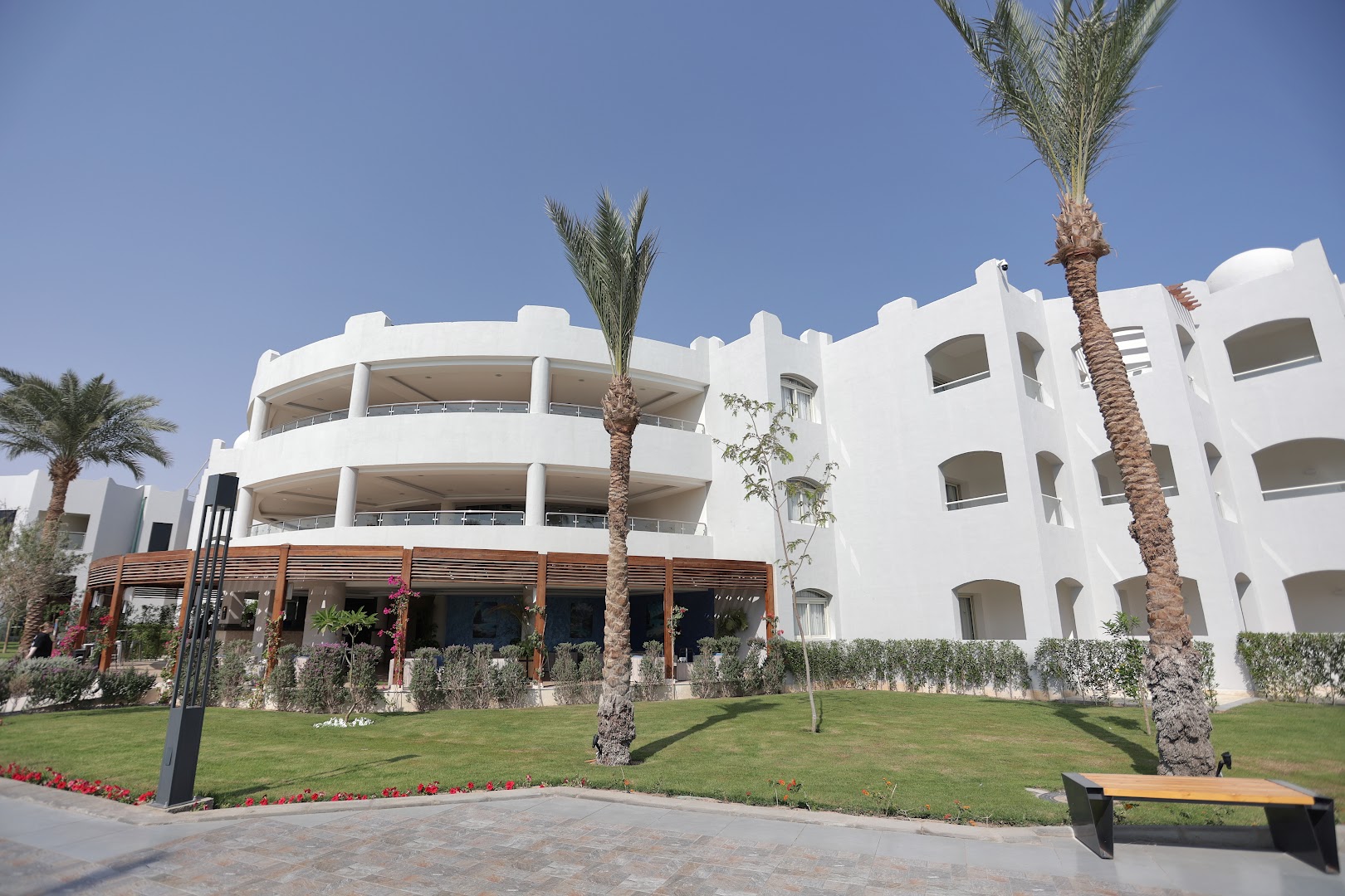 Xanadu Club Makadi Bay gaišā korpusa fasāde ar balkoniem, palmām un atpūtas zonu pirmajā stāvā pie zālāja