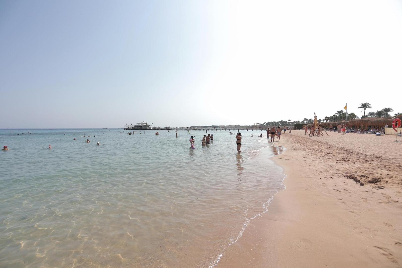 Xanadu Club Makadi Bay smilšu pludmale ar lēzenu ieeju jūrā un atpūtniekiem — dzidrs ūdens un garš koka mols tālumā