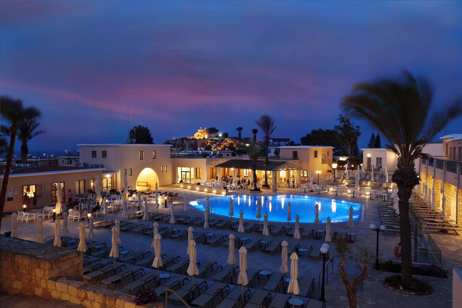 Louis St Elias Resort & Waterpark panorāmas skats vakarā Protarasā — izgaismots galvenais baseins, restorāna zona un baznī...