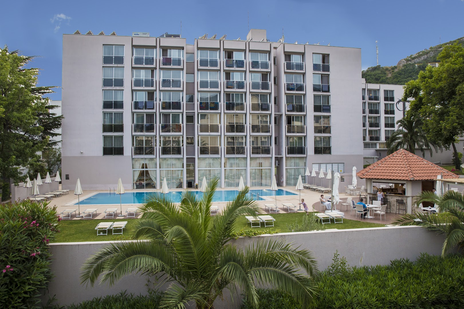 Hotel Tara ēkas fasāde Bečiči kūrortā Melnkalnē ar āra baseinu, saulessargiem un atpūtas zonu zālienā pie viesnīcas korpusa