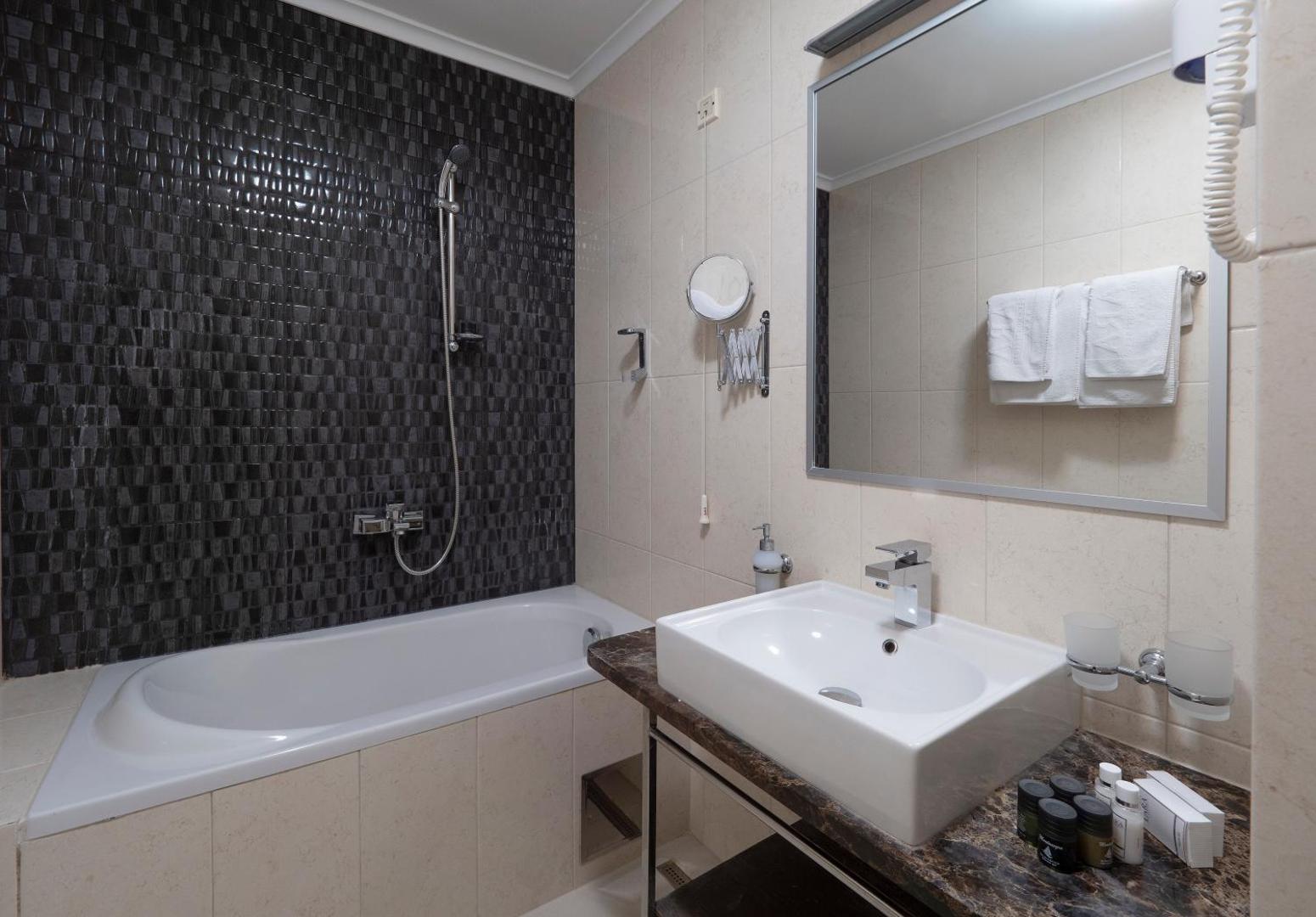 Hotel Tara numura vannas istaba ar vannu, tumšām mozaīkas flīzēm, lielu spoguli un higiēnas piederumiem piekrastes kūrortā Melnkalnē