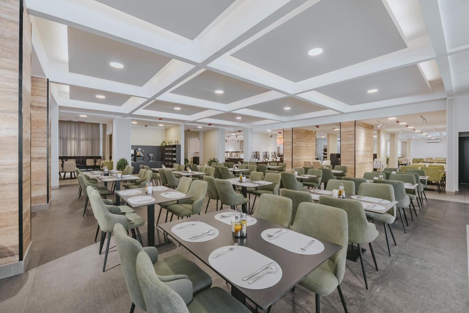 Viesnīcas Hotel Tara restorāna zāle ar pelēkiem krēsliem un saklātiem galdiem — plašs un mūsdienīgs interjers ar ģeometriskiem griestu elementiem