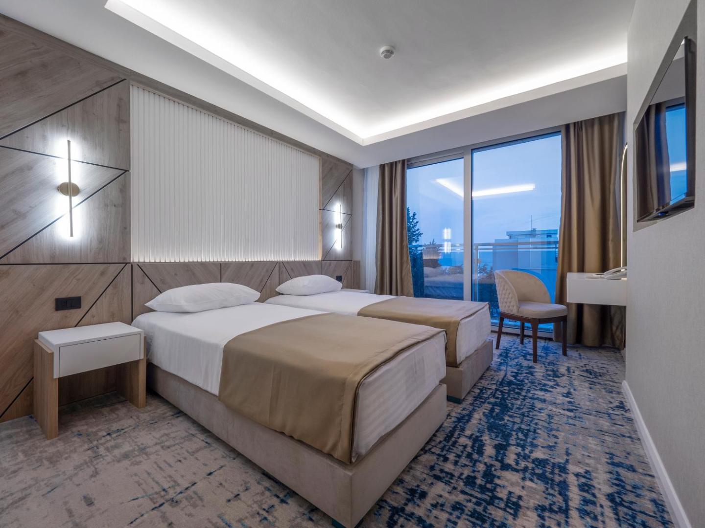 Hotel Tara moderns divvietīgs numurs ar balkonu Bečiči, Melnkalnē — gaišs interjers, koka sienas paneļi un skats uz jūru vakarā